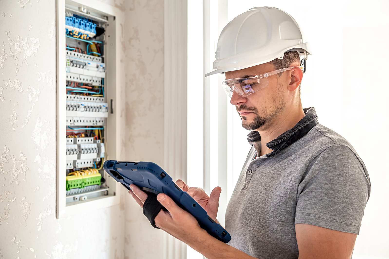 Man, an electrical technician working in a switchboard with fuses, uses a tablet.